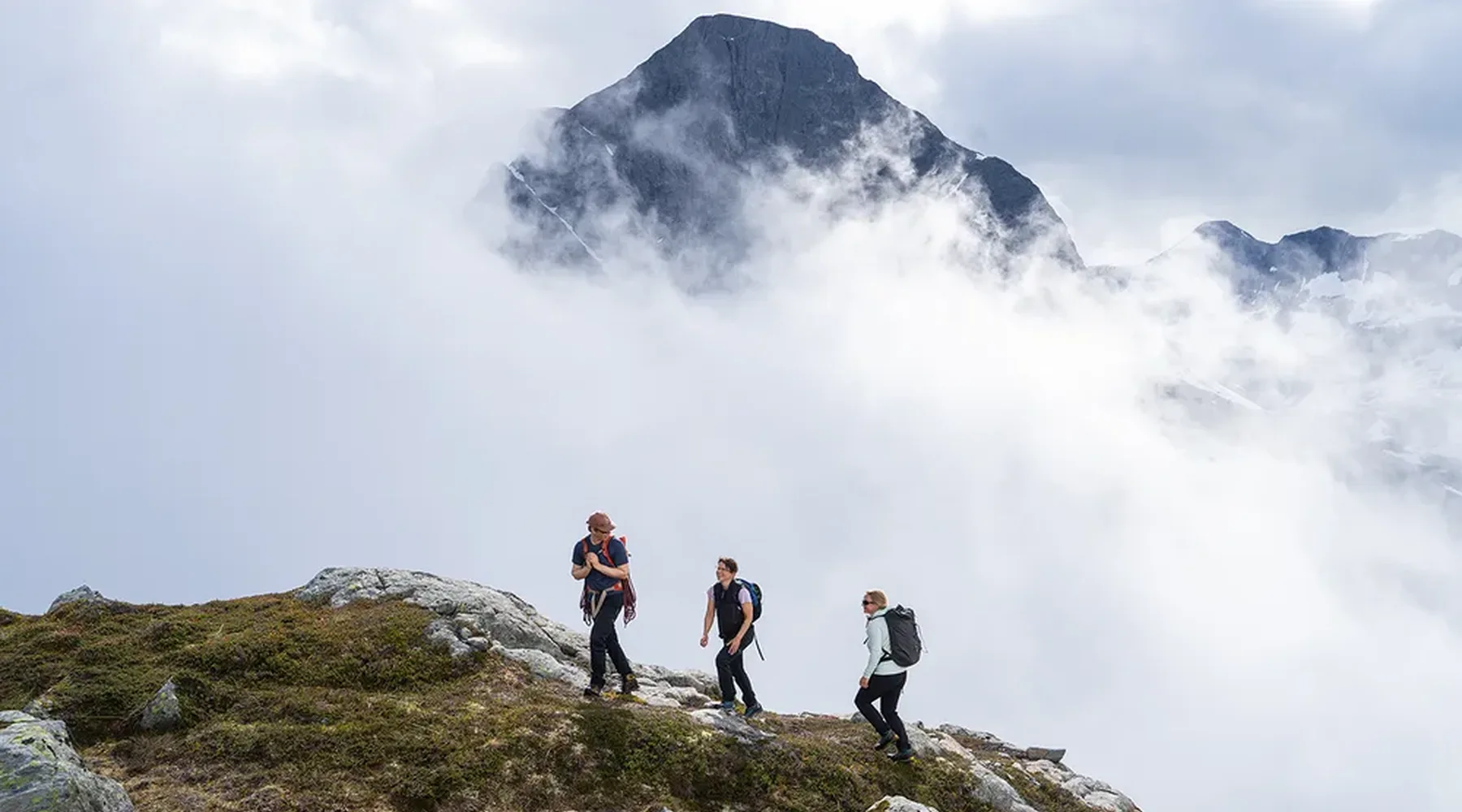 Tre turgåere på fjellrygg med tåke og fjelltopp bak